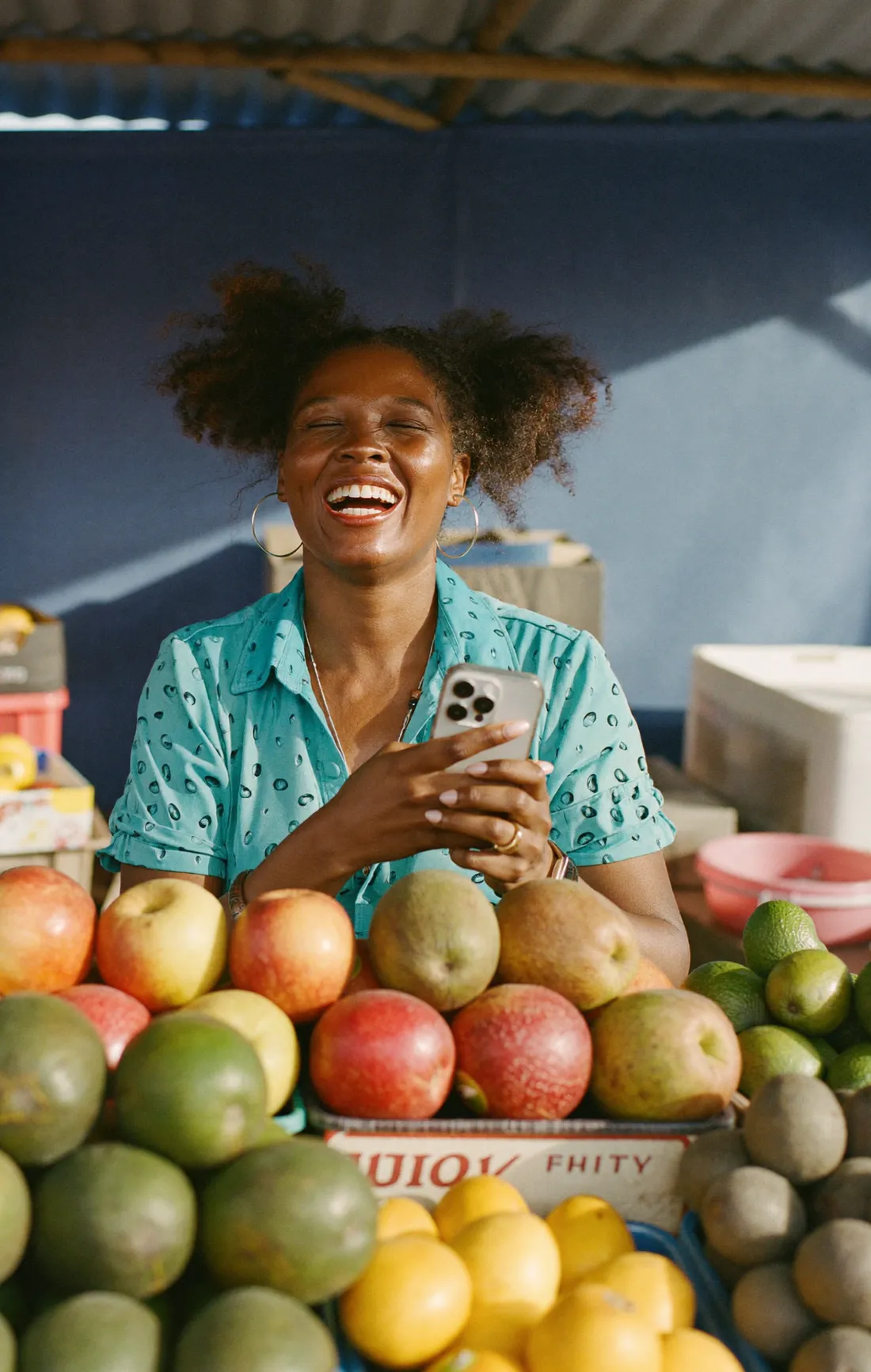 Une marchande haïtienne, souriante, téléphone en main devant son étal de fruits